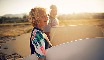 Retired couple walking to ocean with surf boards.